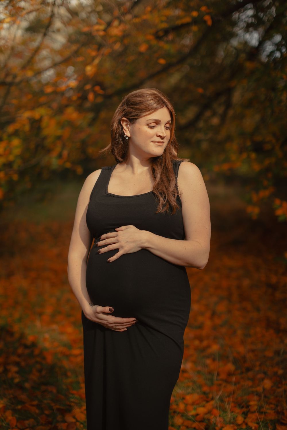 A maternity photo shoot in a fall forest with autumn leaves showing an expectant mother in a black dress.