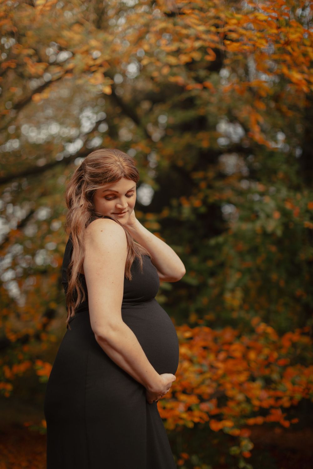 A figure in a black sleeveless dress poses expressively against vibrant autumn foliage in a forest setting.
