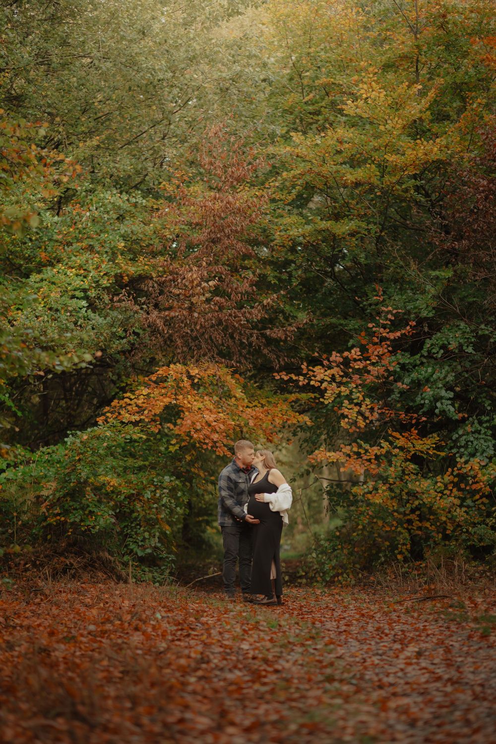 A couple embraces on a path surrounded by vibrant autumn foliage in warm orange and green hues.