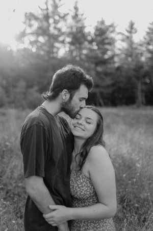 Romantic moment shared in a sunlit field surrounded by tall grass and pine trees.
