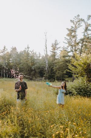 Playful couple dancing through a sunny wildflower field near pine trees.