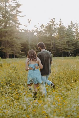 A couple walking through a field of yellow wildflowers at sunset with evergreen trees in the background.