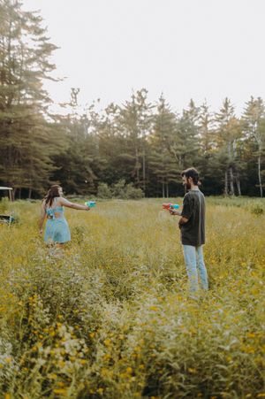 Two people playing with water guns in a meadow filled with yellow flowers and pine trees.