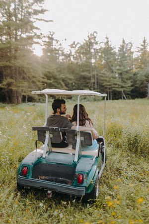 A vintage golf cart drives through a wildflower meadow surrounded by tall evergreen trees.