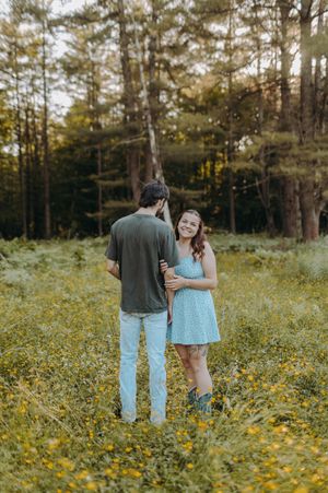 Couple walking through field of yellow wildflowers with pine forest backdrop.