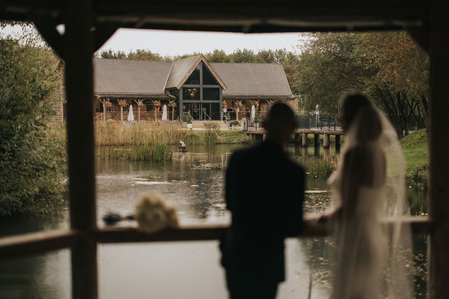 Silhouettes of a wedding couple seen through a covered bridge frame overlooking a rustic lakeside lodge at The Grand Lodge Oaklands.