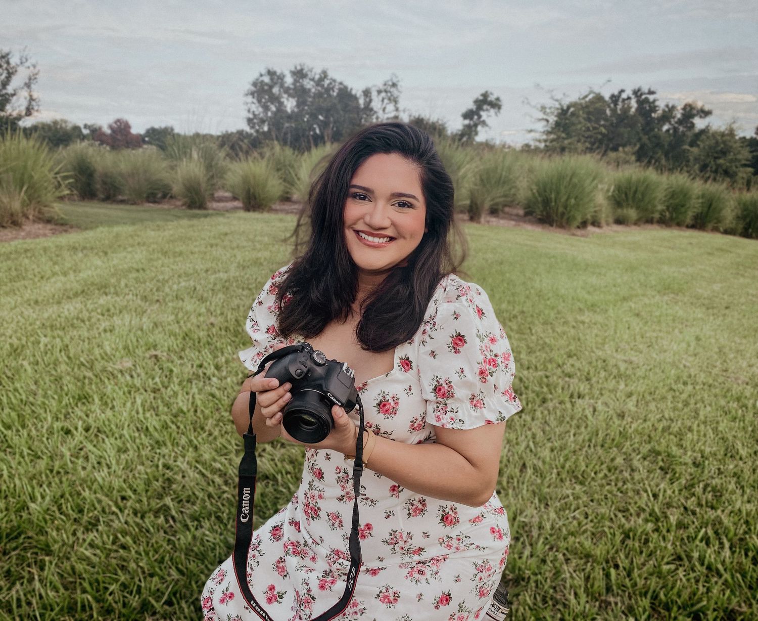 Smiling young woman in floral dress holding a Canon camera while sitting in a green field.