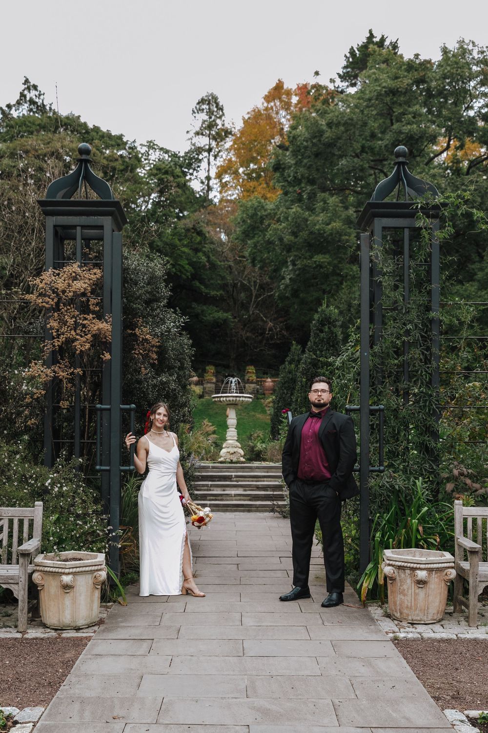 Wedding ceremony setup in a garden with stone pillars, green archways and planters along a paved path.