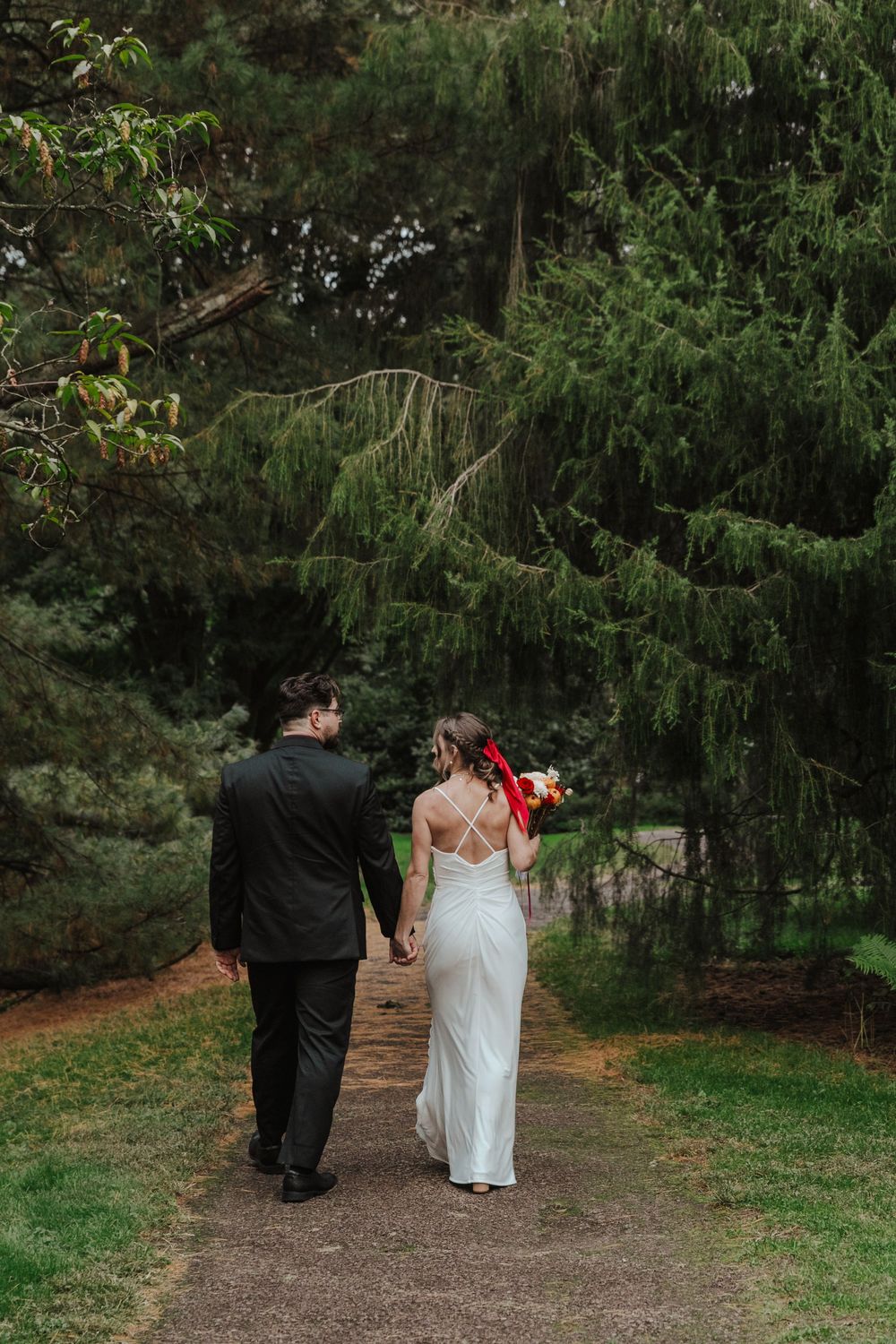 A couple walks hand in hand down a garden path beneath towering evergreen trees at a wedding venue.