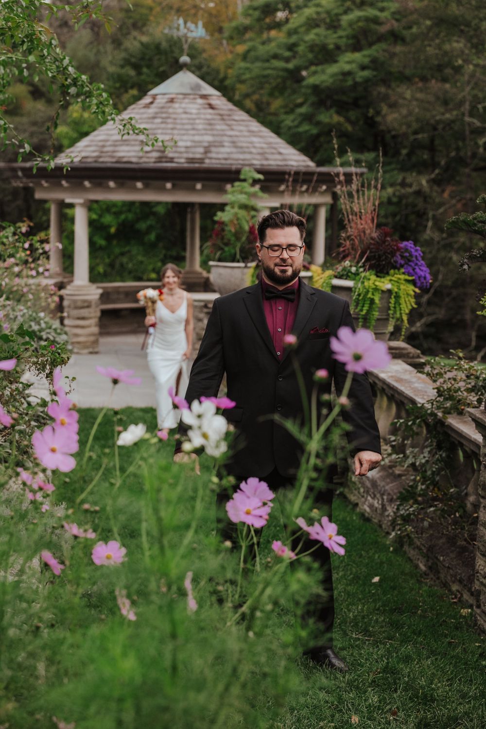 A romantic bride and groom pose in a garden setting with pink cosmos flowers in foreground and gazebos in background.