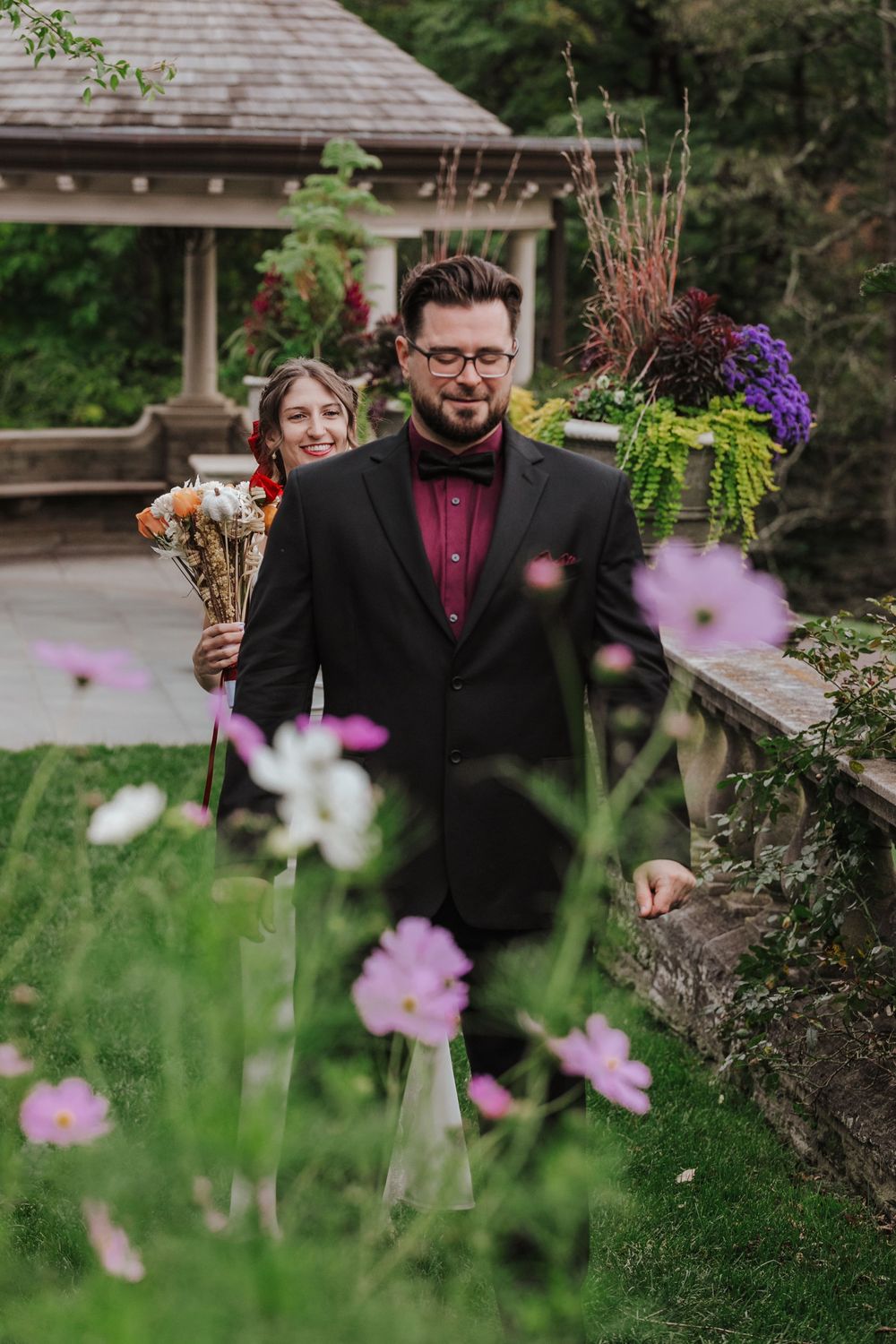 A person in formal black attire walks through a lush garden setting with purple flowers in the foreground.