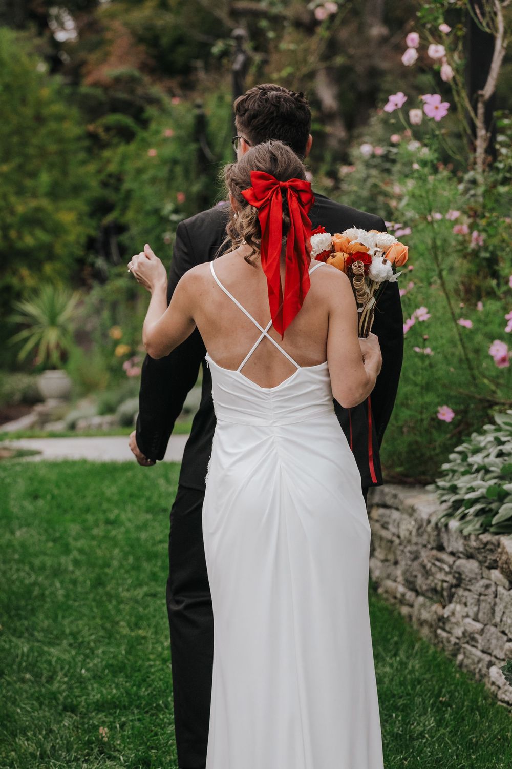 A couple in wedding attire stands in a garden with pink flowers, the bride wearing a white dress with a red bow.