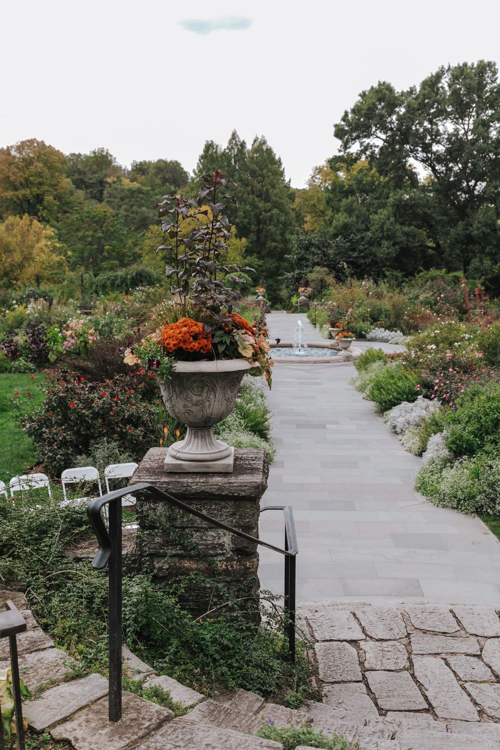 A scenic garden walkway features a series of urns filled with vibrant flowers alongside a curved paved path lined with greenery.
