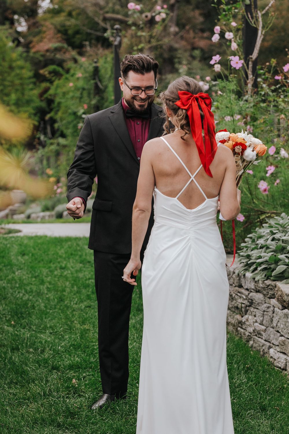 A couple shares a moment in a garden setting, with the bride wearing a white strappy dress and red hair accessory.