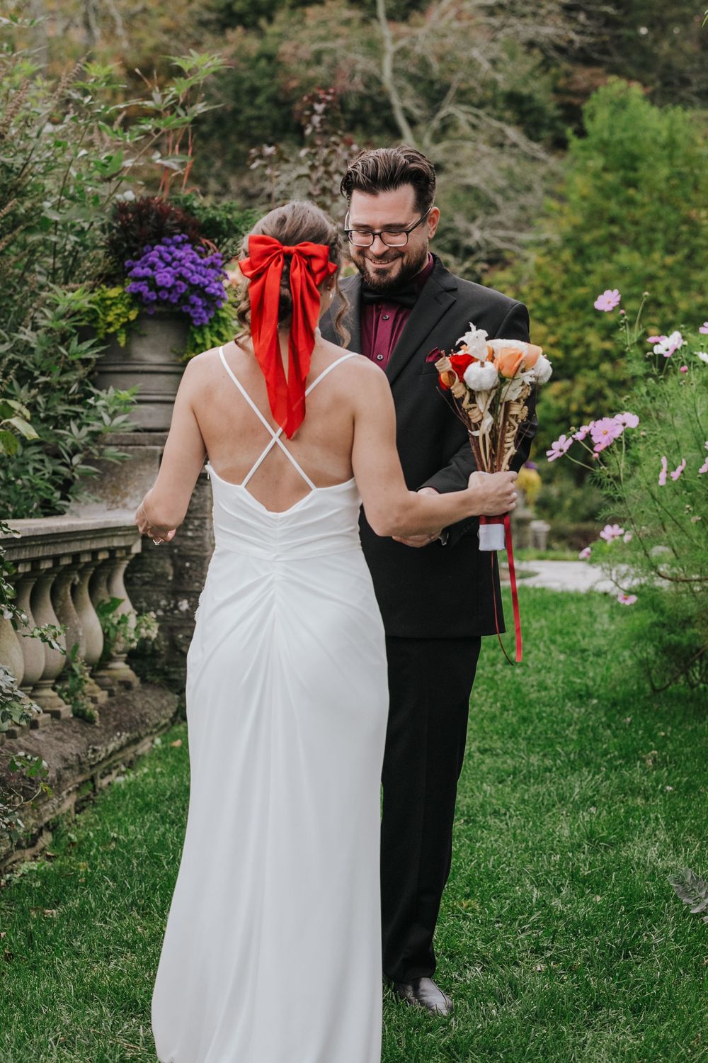 A couple shares a candid moment in a garden with the bride in white dress and red hair ribbon holding a bouquet.