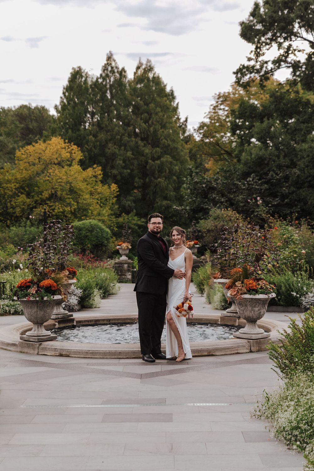 Wedding couple dancing together on an outdoor stone patio with autumn flowers and decorative urns in elegant garden setting.