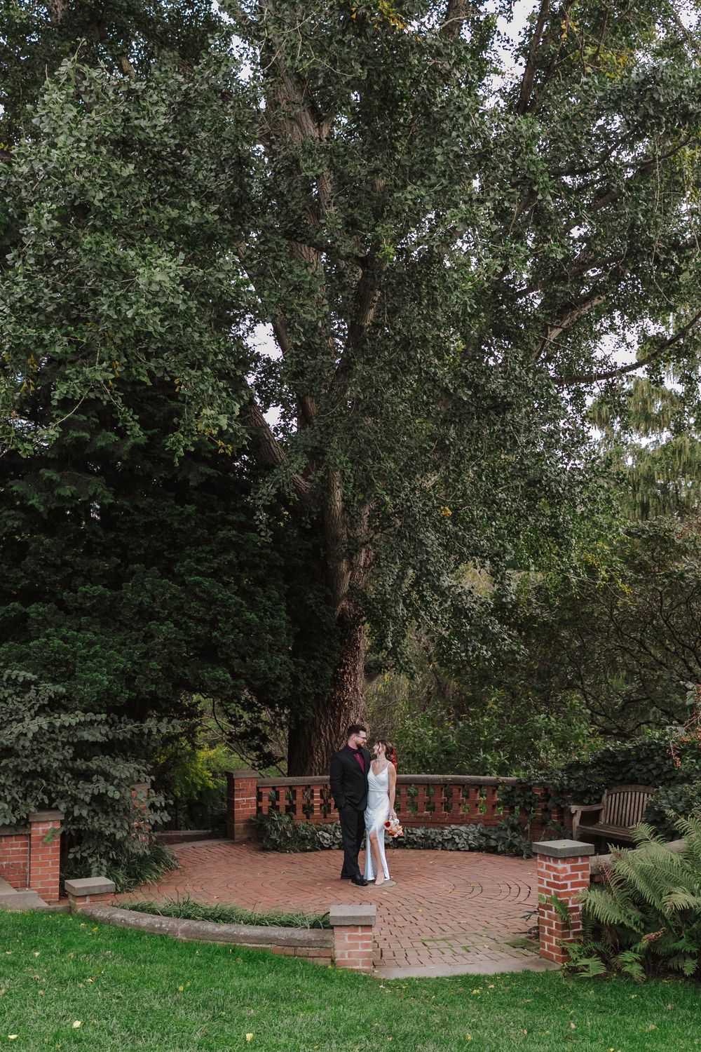 An elegant couple stands together on a circular brick patio surrounded by lush greenery and tall trees in a garden setting.