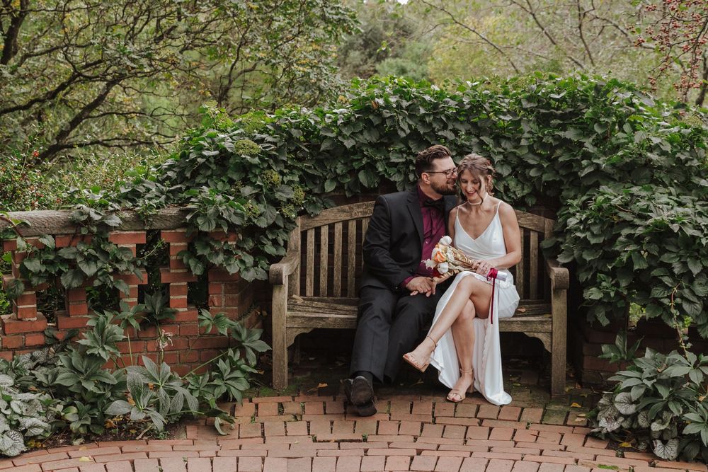 A couple shares an intimate moment on a wooden bench surrounded by lush green foliage in a garden setting.