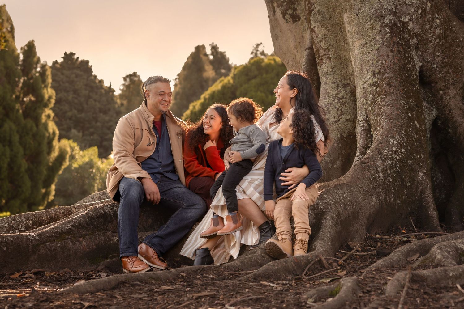 Lifestyle Family portrait of a young family laughing candidly sitting at the bottom of a large tree with golden sun rays