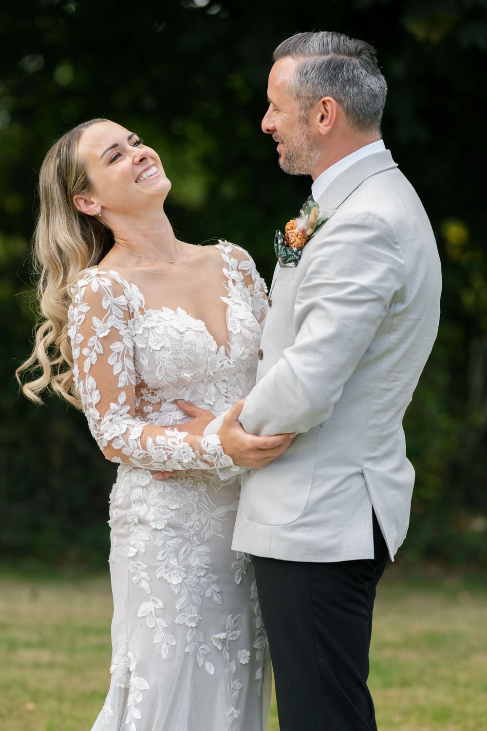 Newlywed couple shares a joyful moment outdoors, with bride in lace dress and groom in light gray jacket.