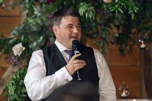 Wedding guest in a formal vest and white shirt raises a glass while giving a toast at a reception with greenery decorations.