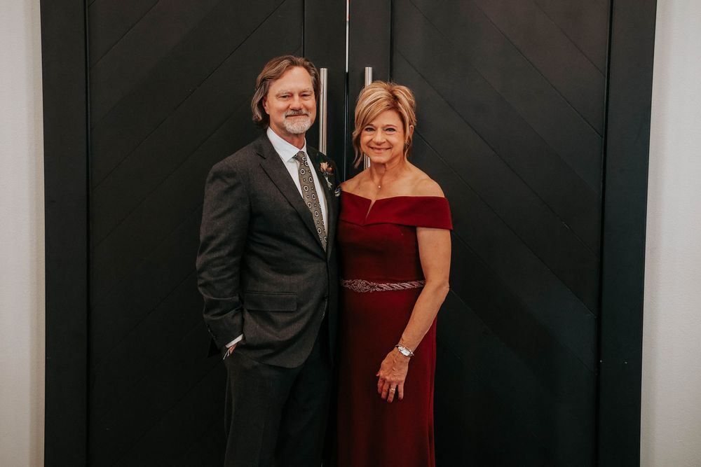 A couple poses together against a dark wall, with him in a black suit and her wearing an elegant burgundy evening gown.