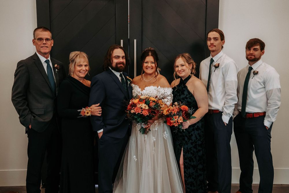 A formal wedding party in elegant black and navy attire poses together against a neutral wall for a group photo.