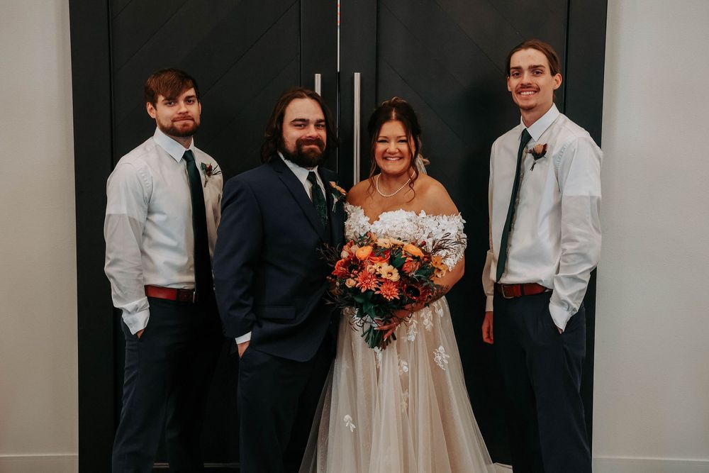 Wedding party group poses formally against a black wall in suits and gowns with orange floral accents.