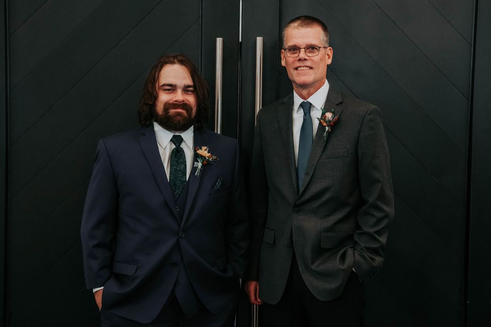 Groomsmen in dark suits standing against black wall at formal wedding event.