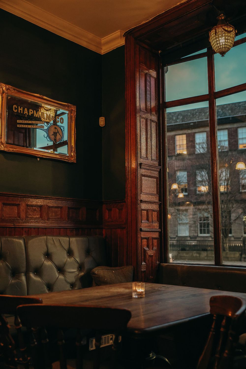 Cozy pub corner with dark wood paneling, leather seating, and large window showing urban buildings.