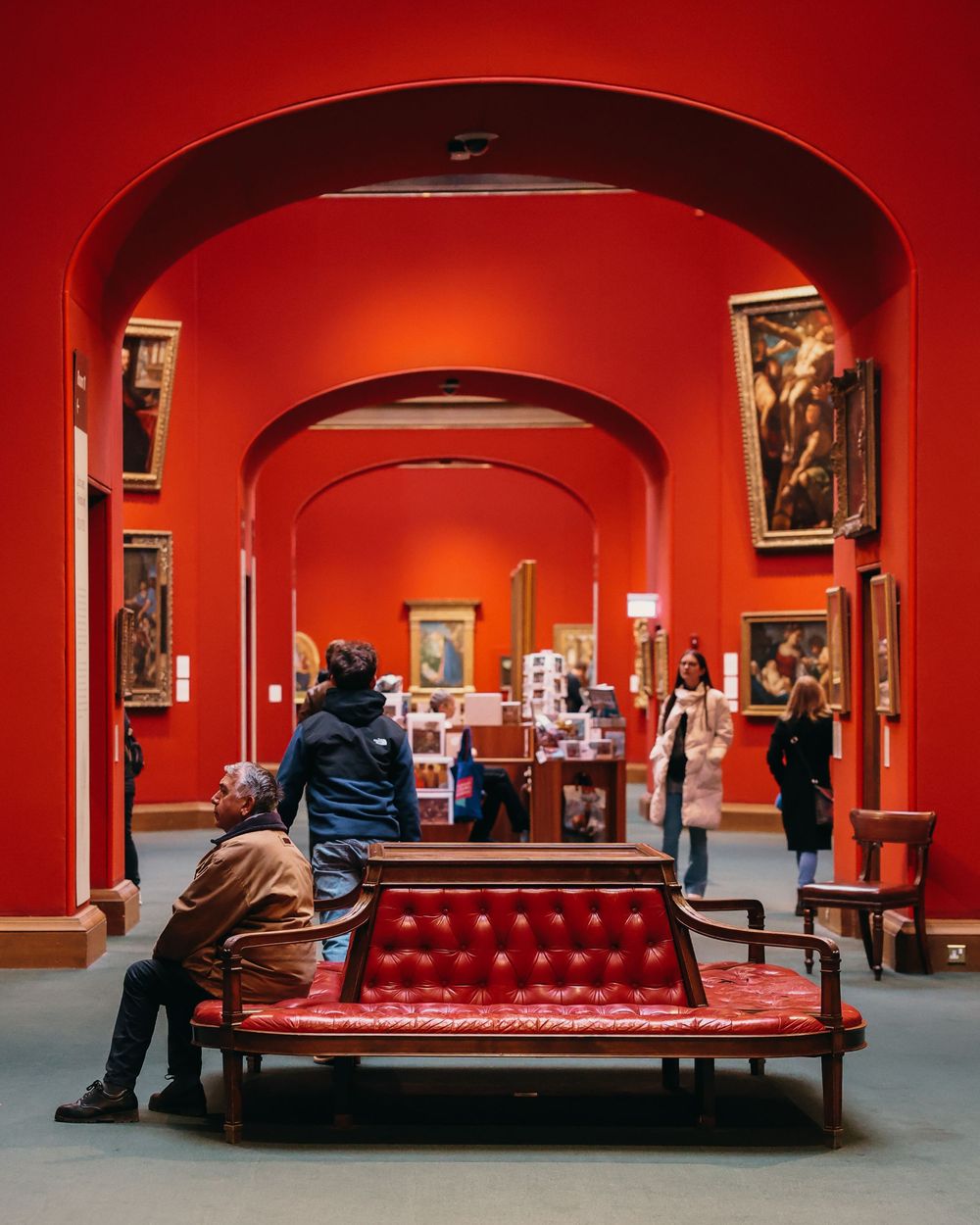 Art gallery interior with red walls, arched doorways, and a tufted red bench in the center.