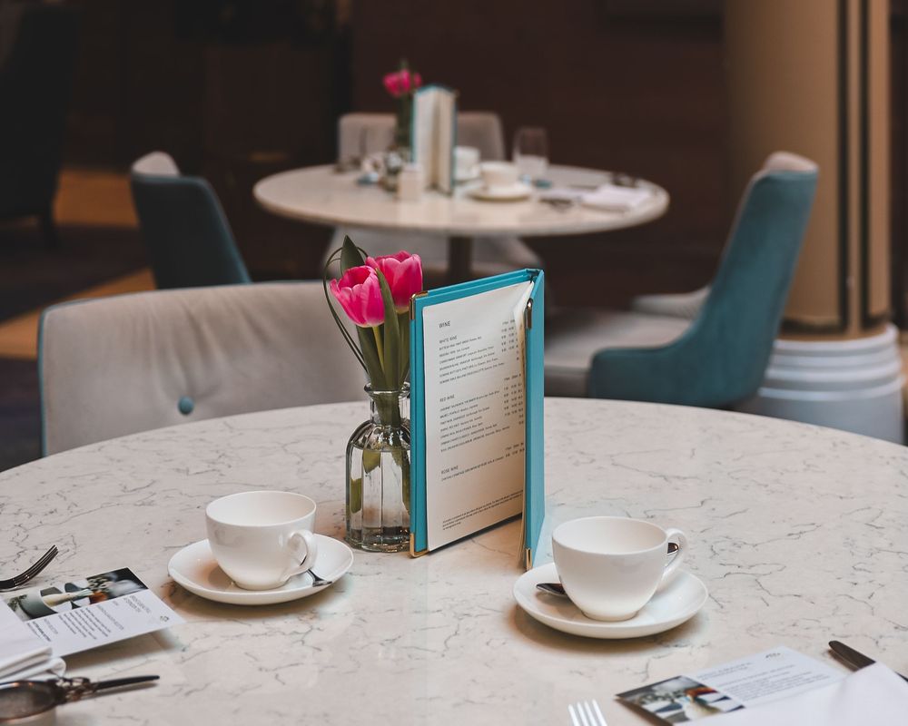 Table setting with two coffee cups and a teal menu alongside pink tulips in a vase on a marble surface.