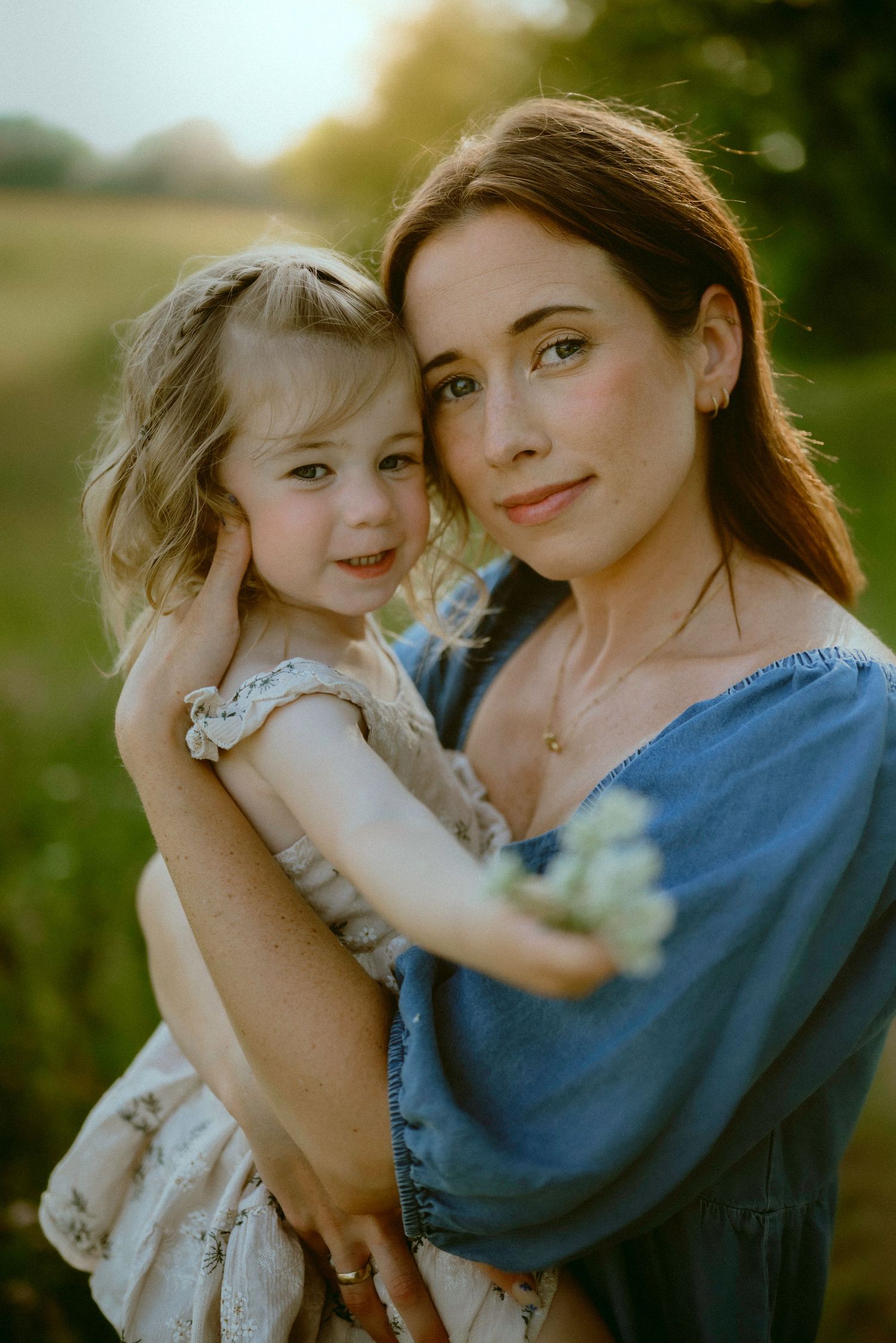 Sun-drenched portrait of parent and child embracing in a meadow at golden hour wearing blue and floral outfits.