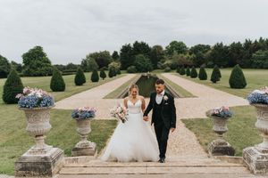 A couple walks down a grand garden path lined with manicured topiary and classical stone urns filled with hydrangeas.