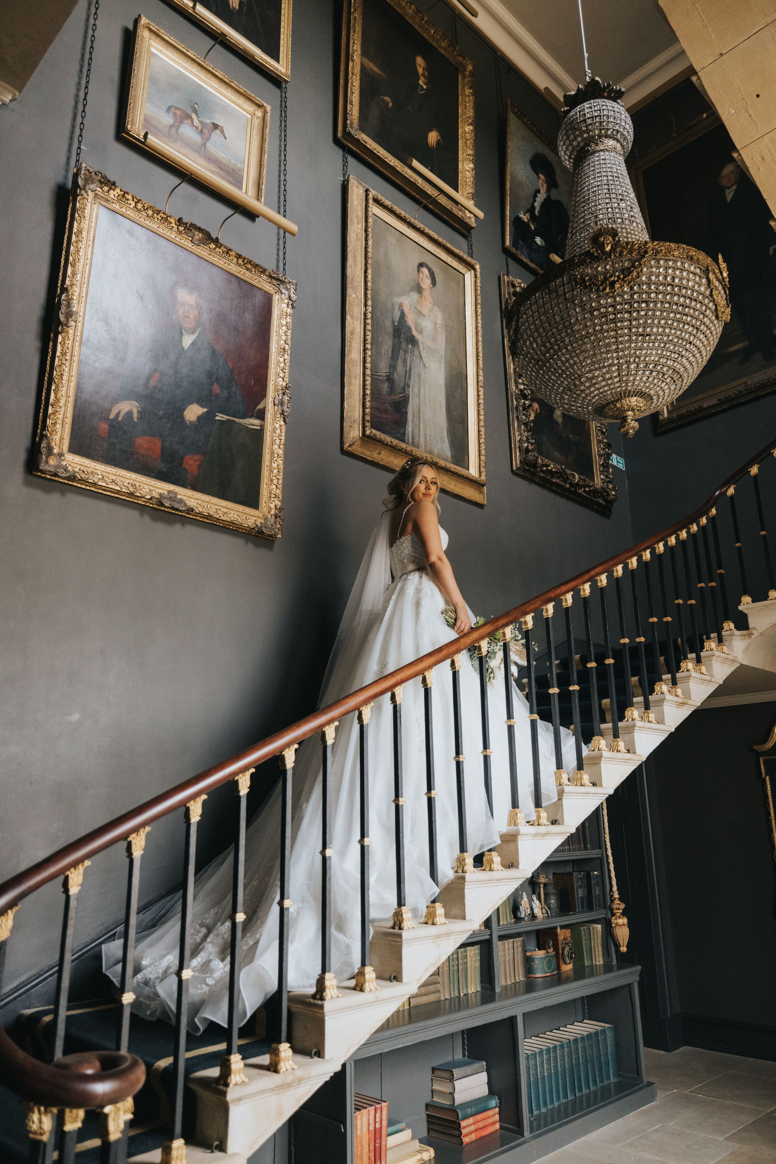 Crystal chandeliers hang above a classic staircase with dark walls and framed artwork in an elegant hallway.
