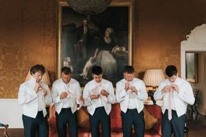 Row of groomsmen in formal attire seated with hands covering their faces in matching poses.