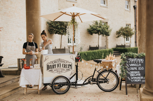 A vintage-style ice cream cart with umbrella and chalkboard sign stands outside a building entrance.