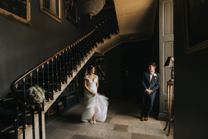 Two people in formal attire stand in an elegant dark interior with a grand curved staircase and dramatic lighting.
