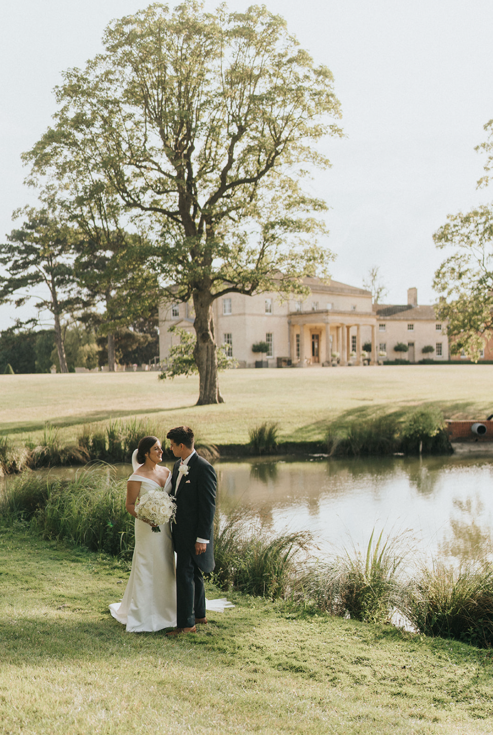 Wedding couple stands near a pond with a grand estate house and tall tree in the background on an elegant lawn.
