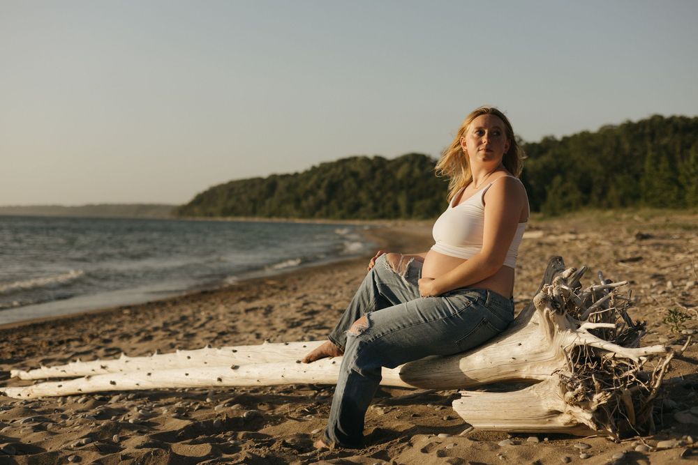 Person sits peacefully on driftwood at sunset on a rocky beach with forested coastline in background.