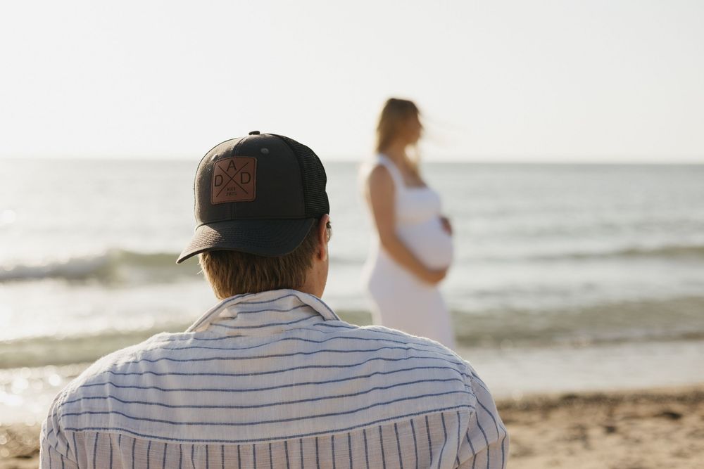 A person in a striped shirt watches someone standing by the ocean at sunset during a beach photoshoot.