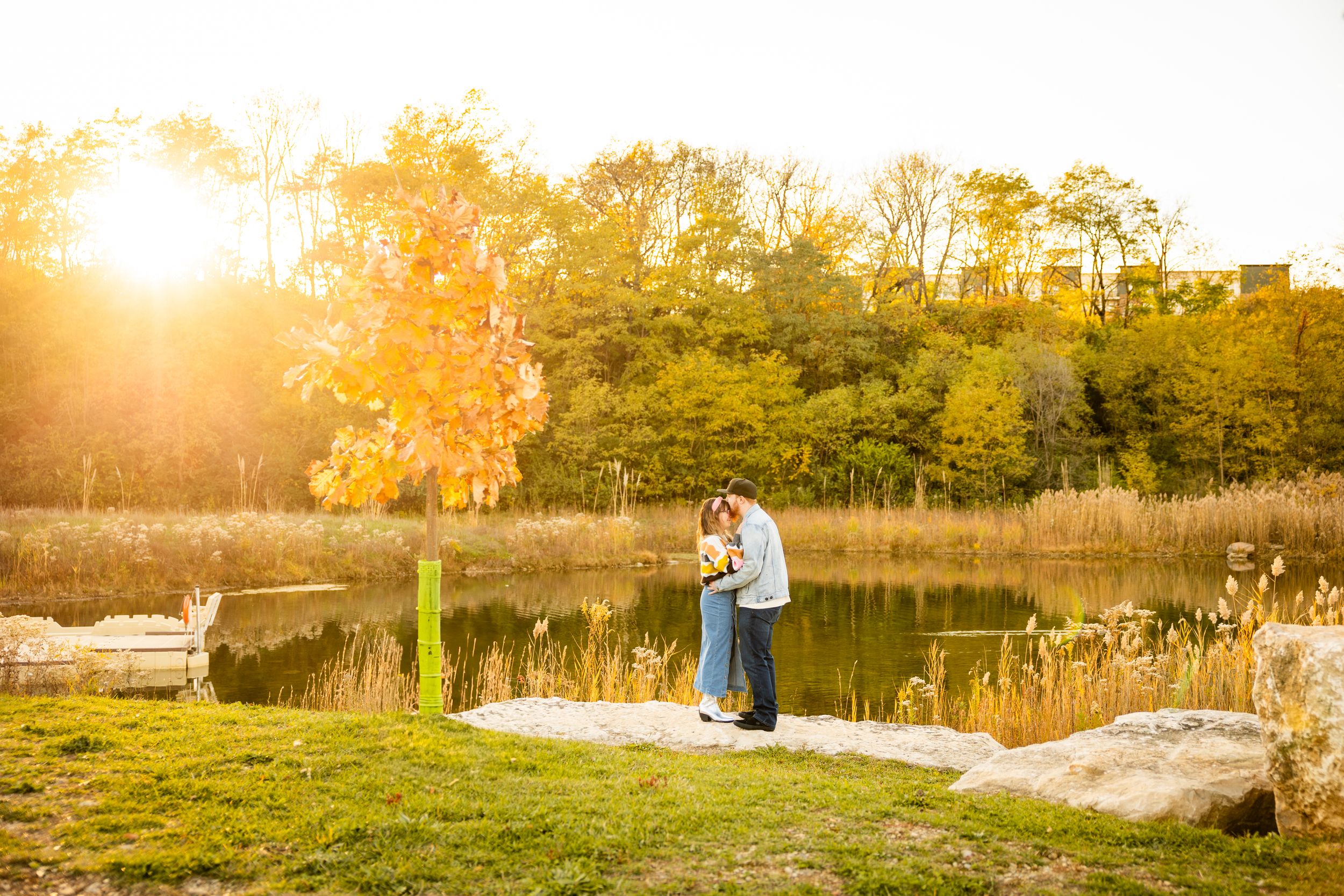 A couple embraces in a golden field during sunset as warm sunlight filters through tall grass and trees.