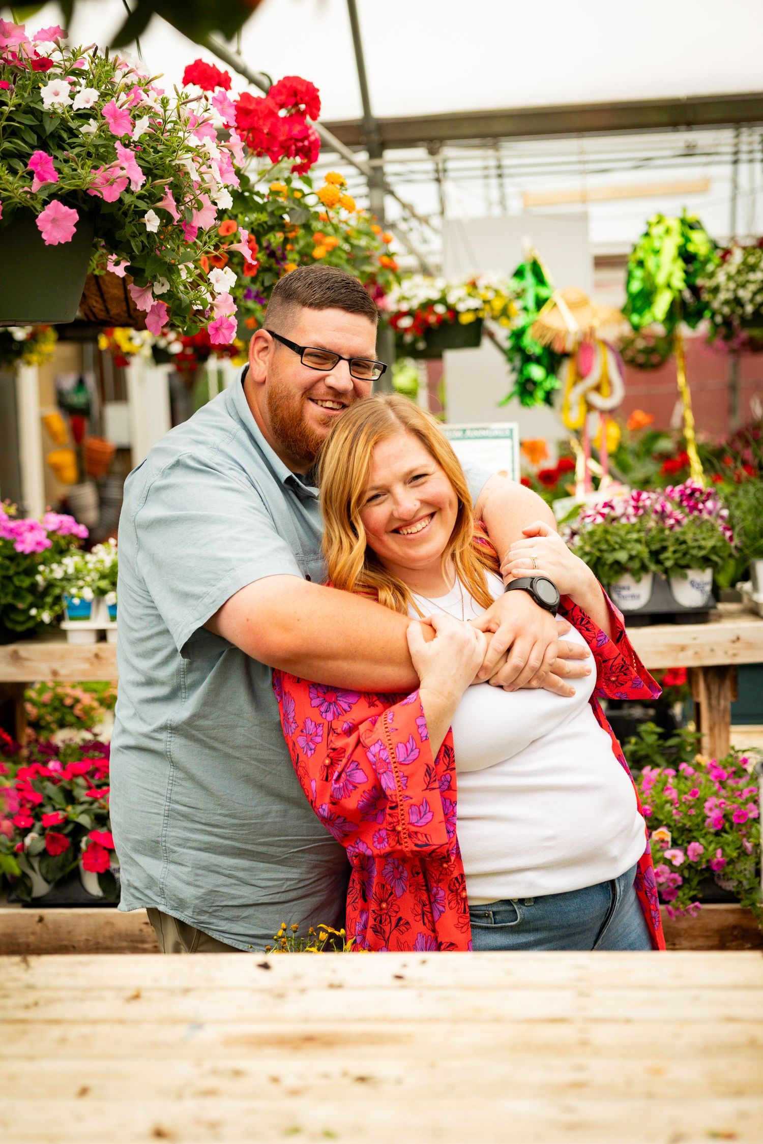 Two shoppers embrace while browsing colorful hanging flower baskets at an outdoor garden center.