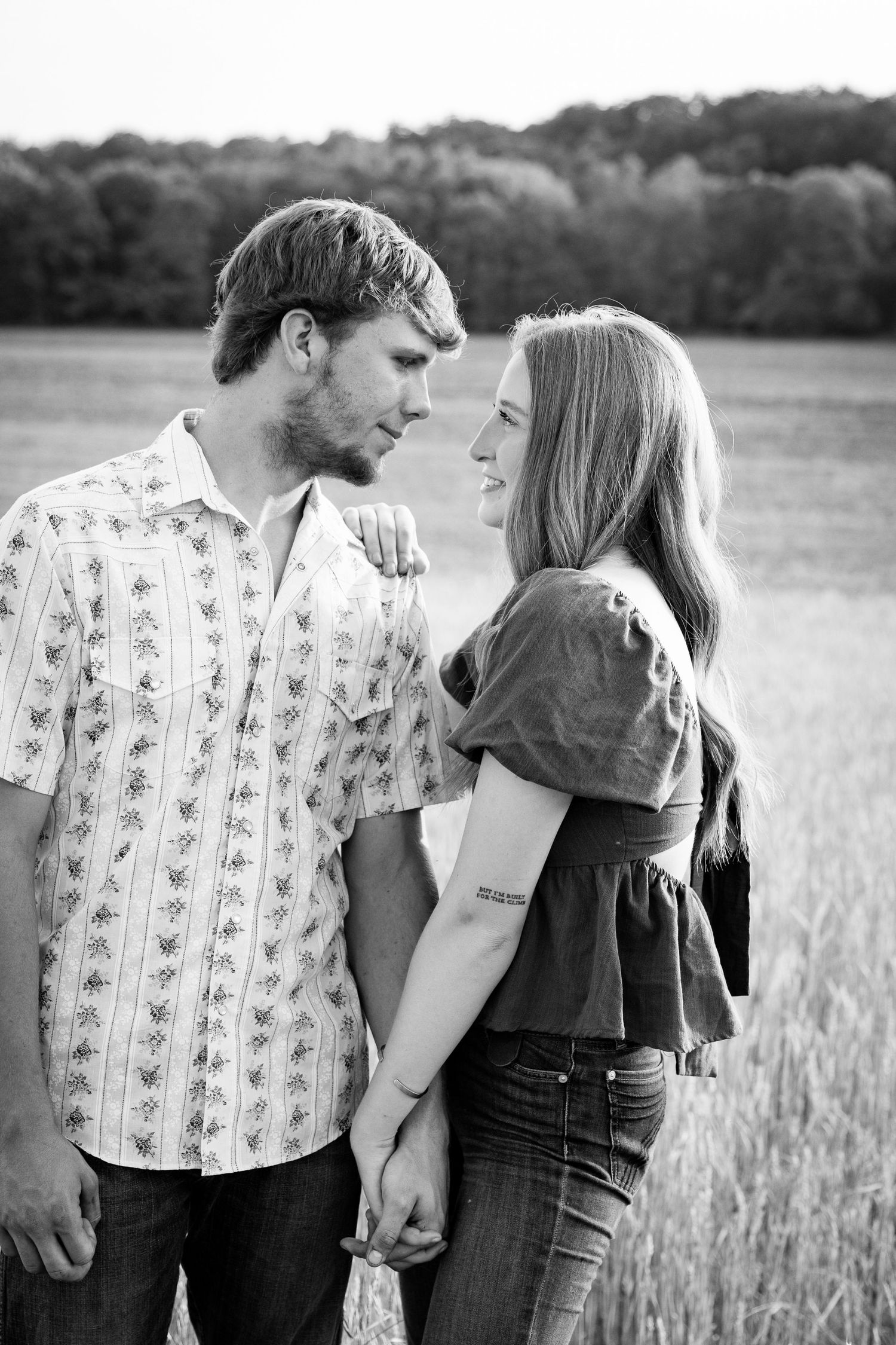 Couple in casual attire standing close together in a sunny wheat field showing romantic connection in black and white.
