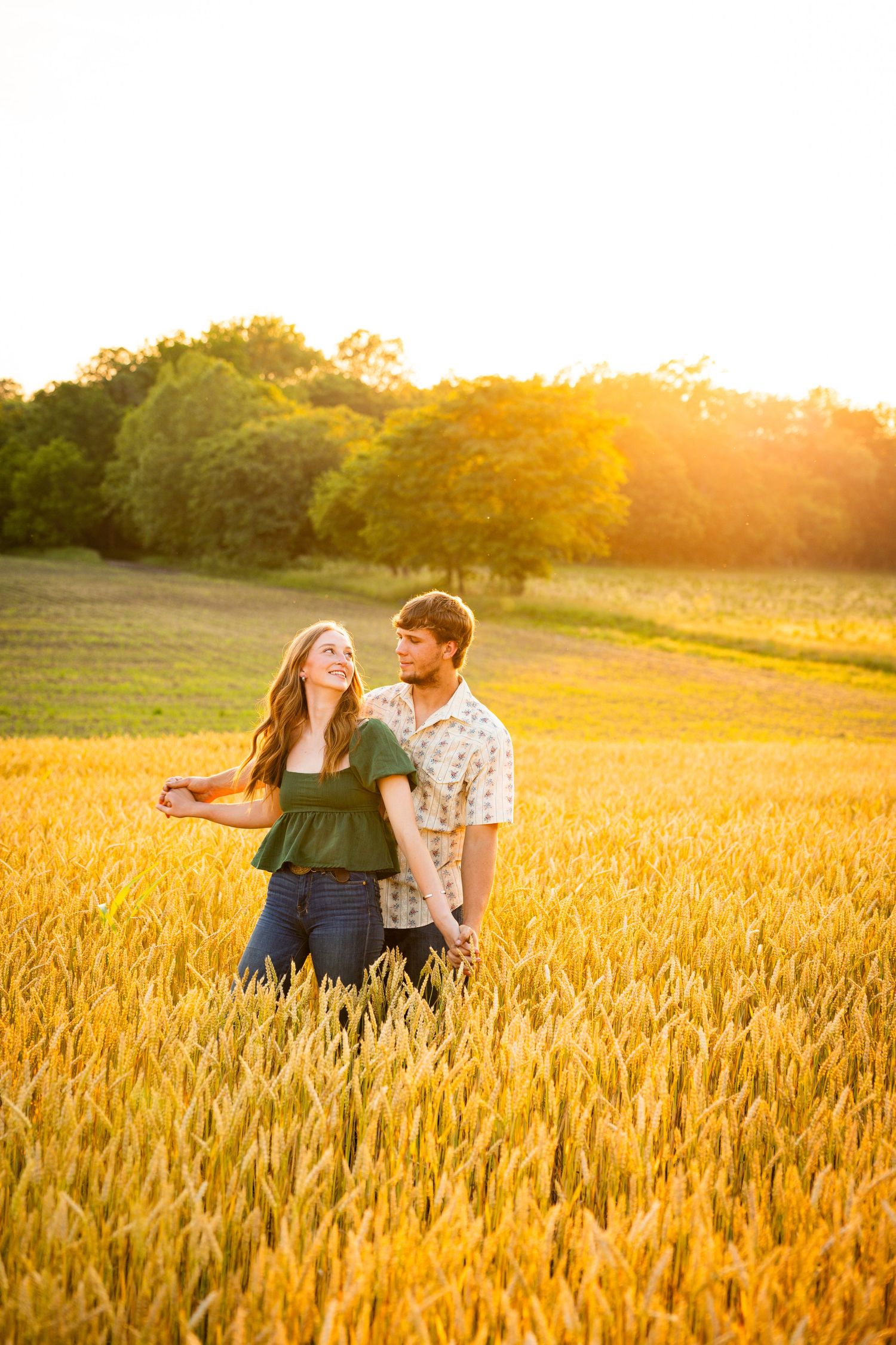 A couple sits together in a golden wheat field during sunset, creating a romantic silhouette against the warm sunlight.