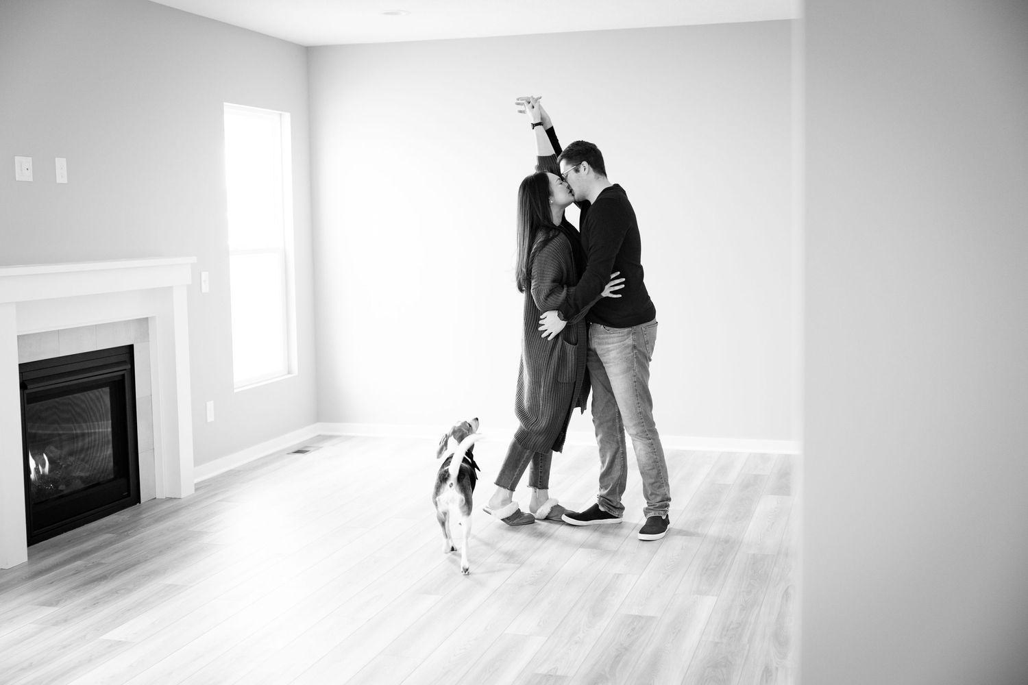 Black and white photo of a couple dancing in an empty room with a small dog by their feet and fireplace visible.