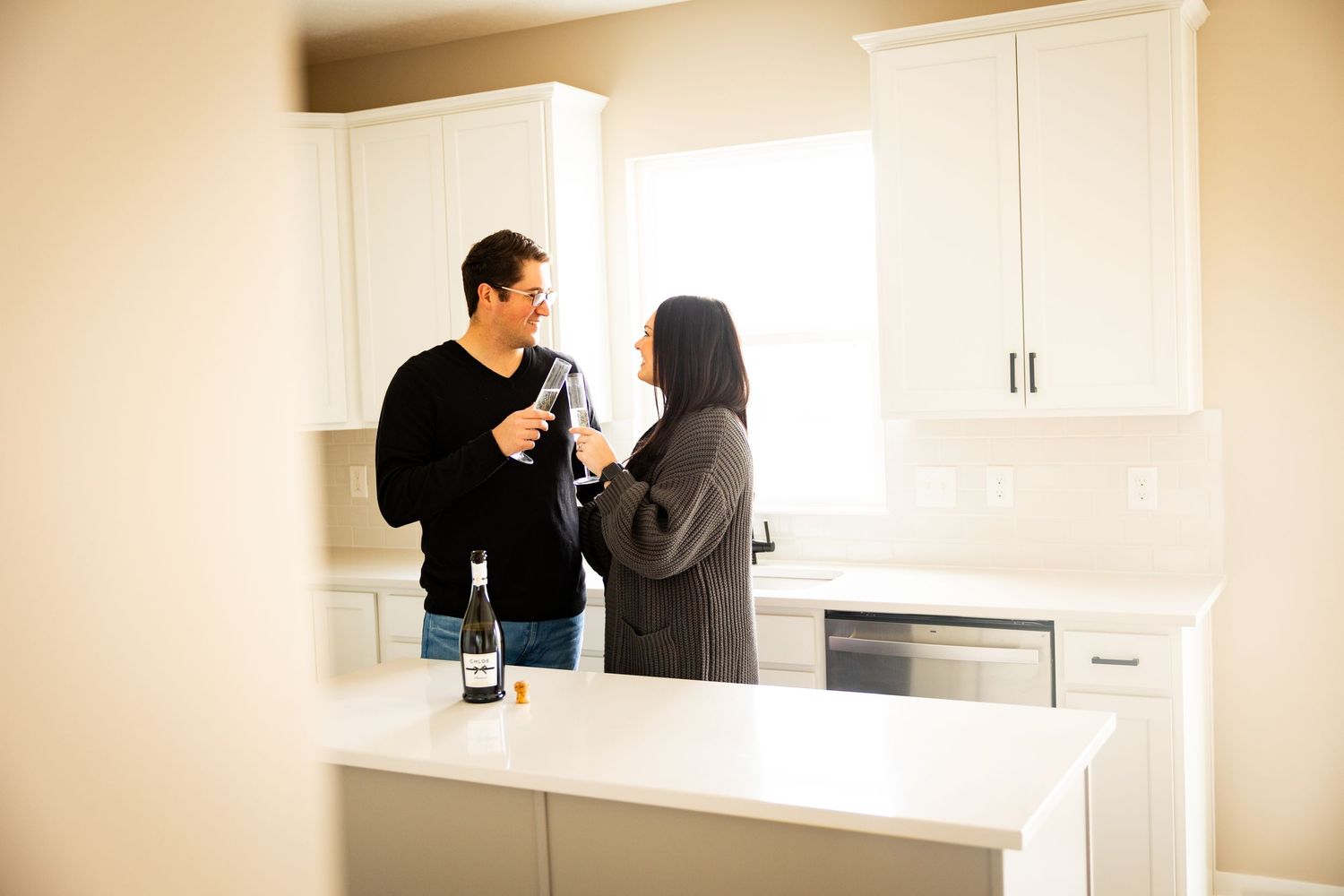 Two people stand and talk while holding wine glasses in a modern white kitchen with quartz countertops.