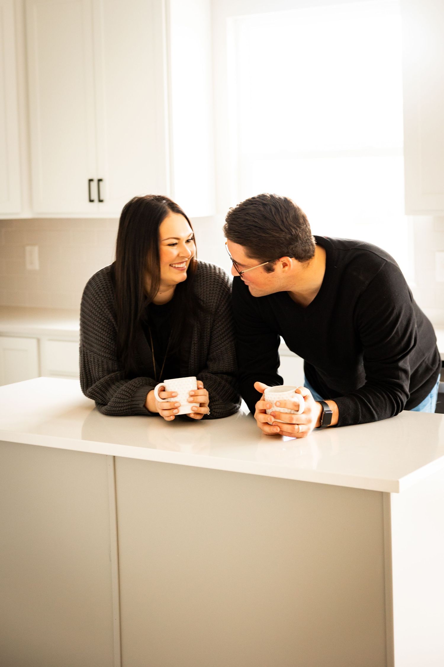 Two people in black clothing lay on a white surface having an intimate conversation over coffee cups.