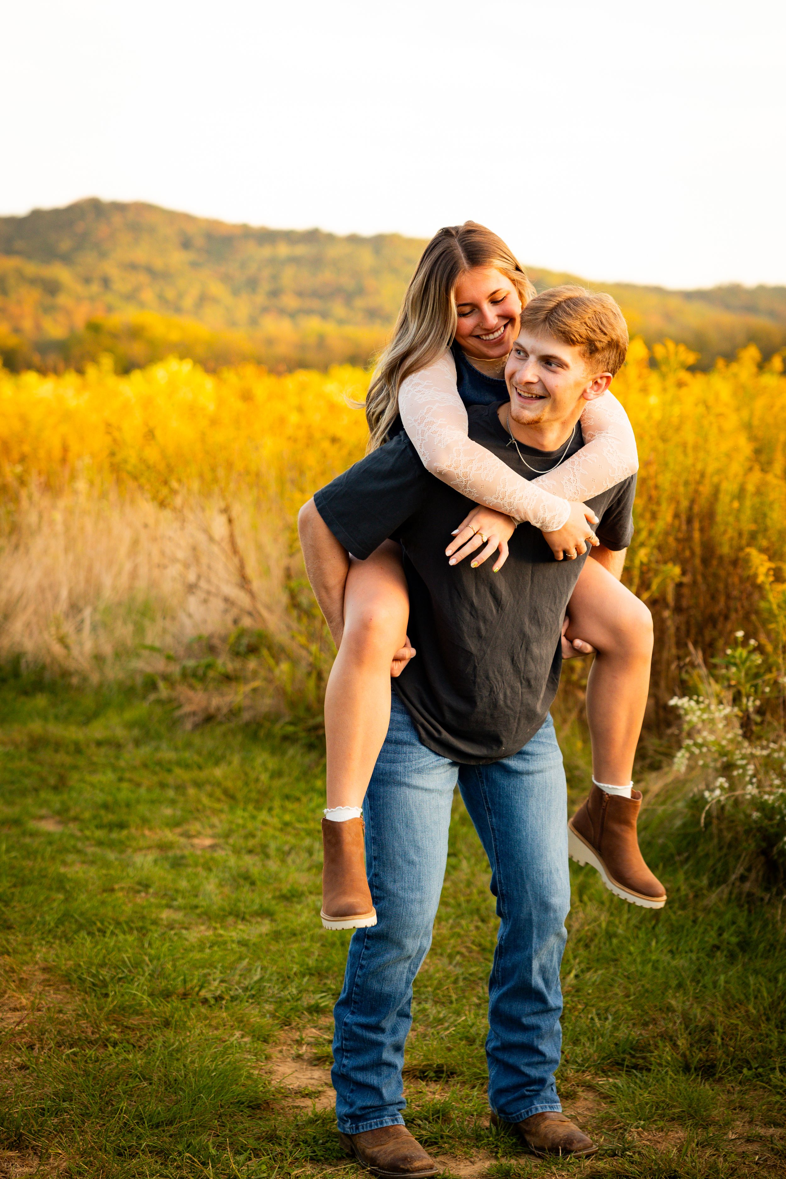 A couple shares a joyful piggyback ride moment in a golden autumn field with hills in the background.