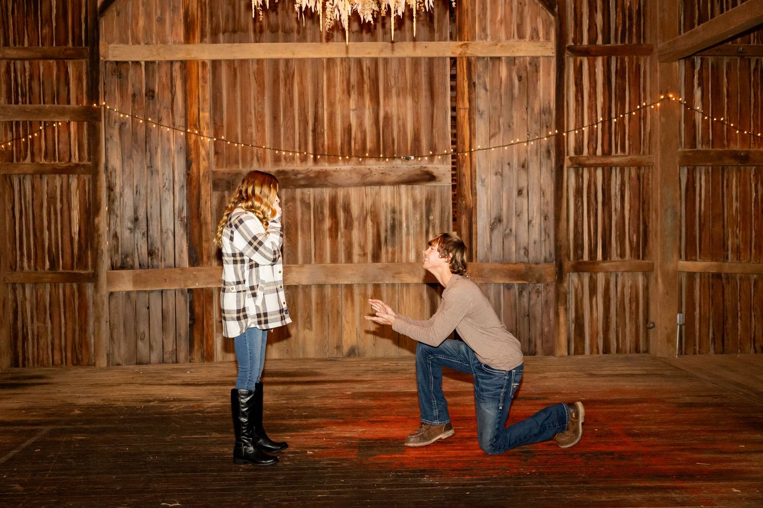 A rustic scene inside a wooden barn with string lights and two people in casual attire standing close together.